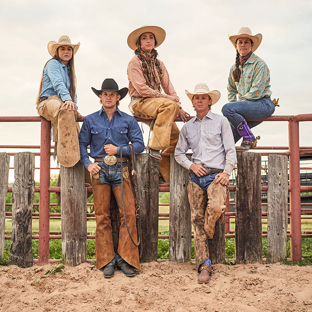 Three women and two men at a ranch wearing jeans, a shirt, cowboy hat, and cowboy boots.